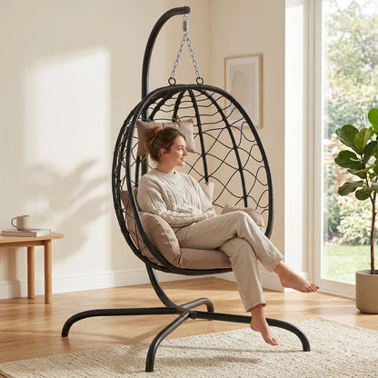Fauteuil suspendu en rotin noir avec coussins beige, vue latérale sur piètement courbé.