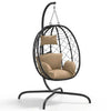 Fauteuil suspendu en rotin noir, coussins beige et structure ovale sur piètement courbé.